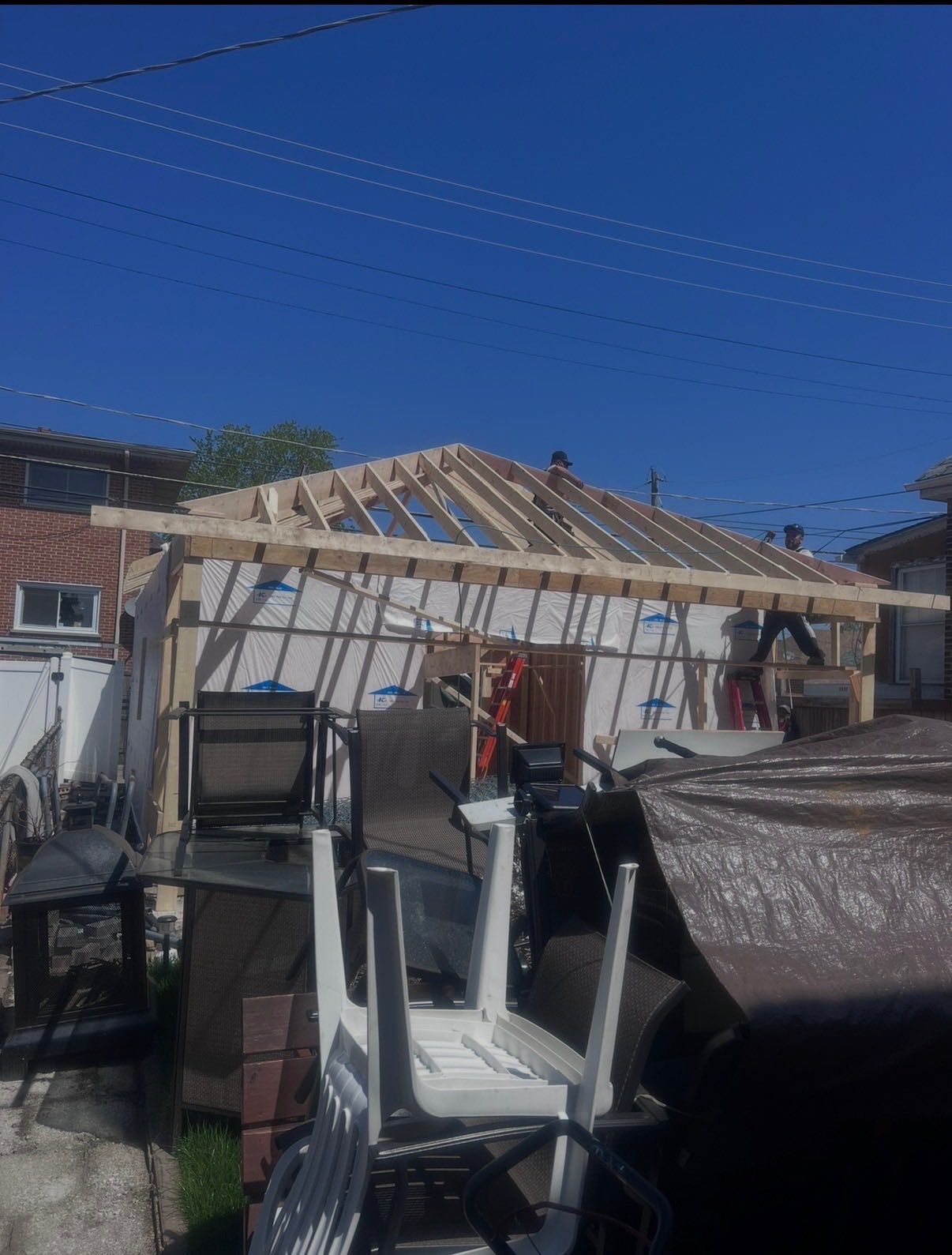 Workers build a wooden roof frame over the structure of a house extension in a residential backyard on a sunny day.