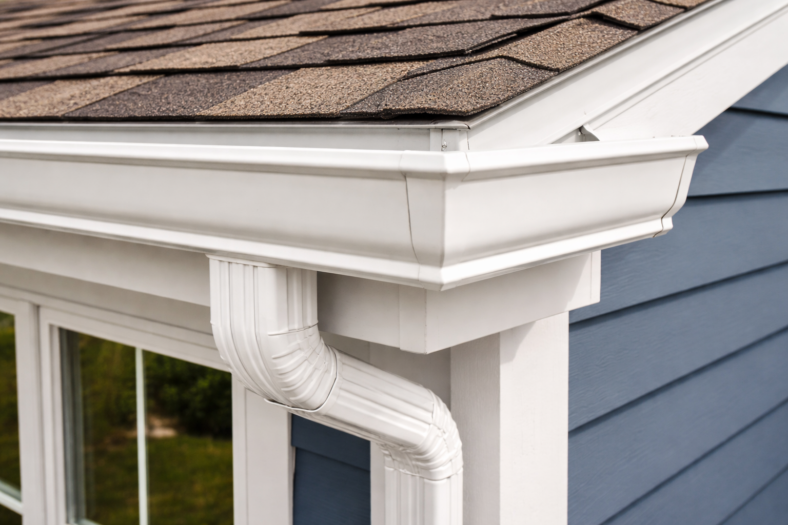 Close-up of a white gutter and downspout attached to the corner of a house with blue siding and brown roof shingles.