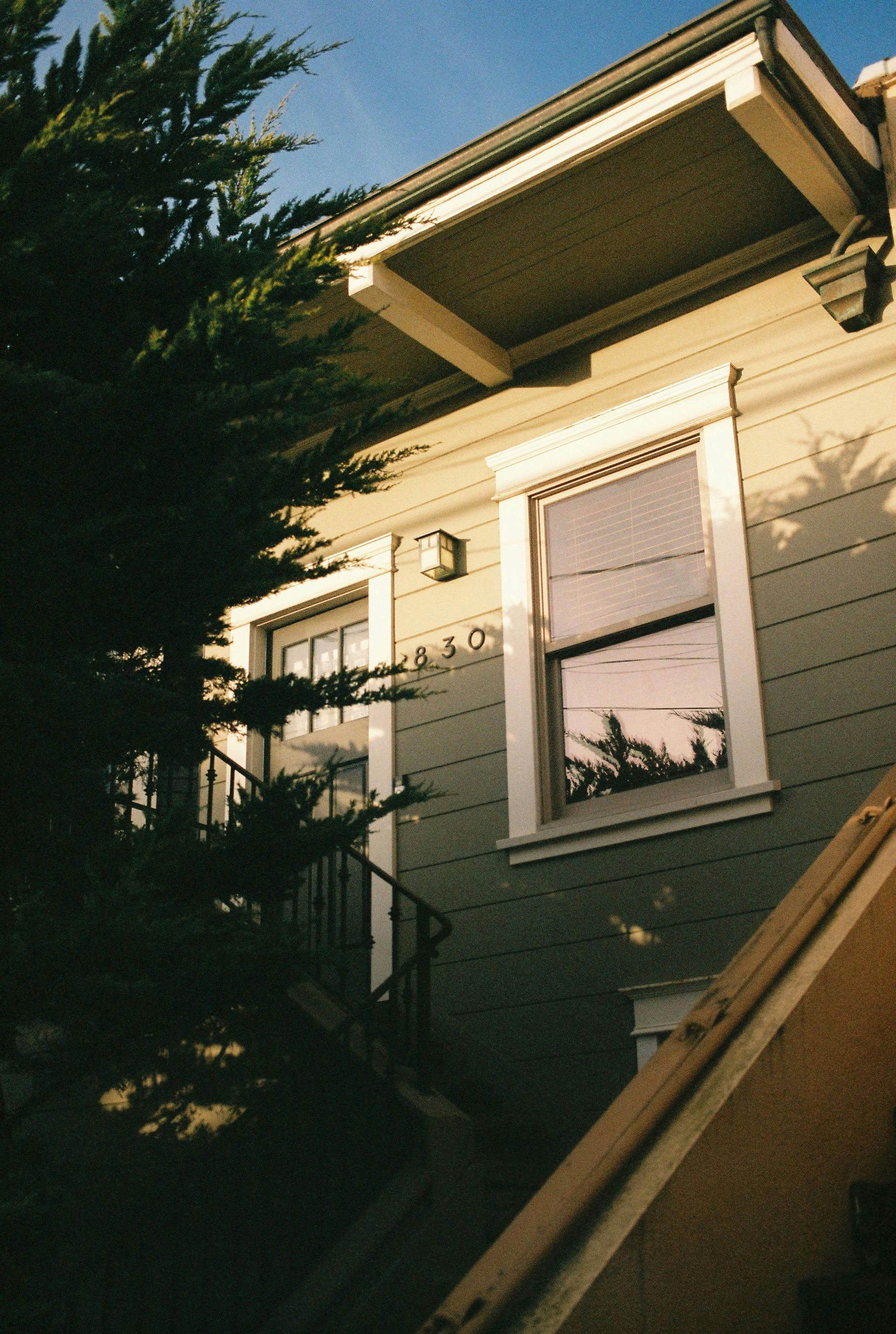 A beige house exterior with a window, a partial view of an entrance with stairs, and a large tree in the foreground.