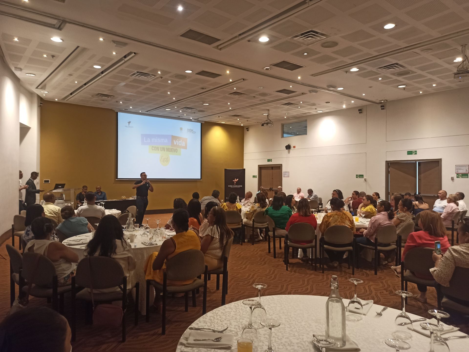 Un grupo de personas están sentadas en mesas en una sala grande viendo una presentación.