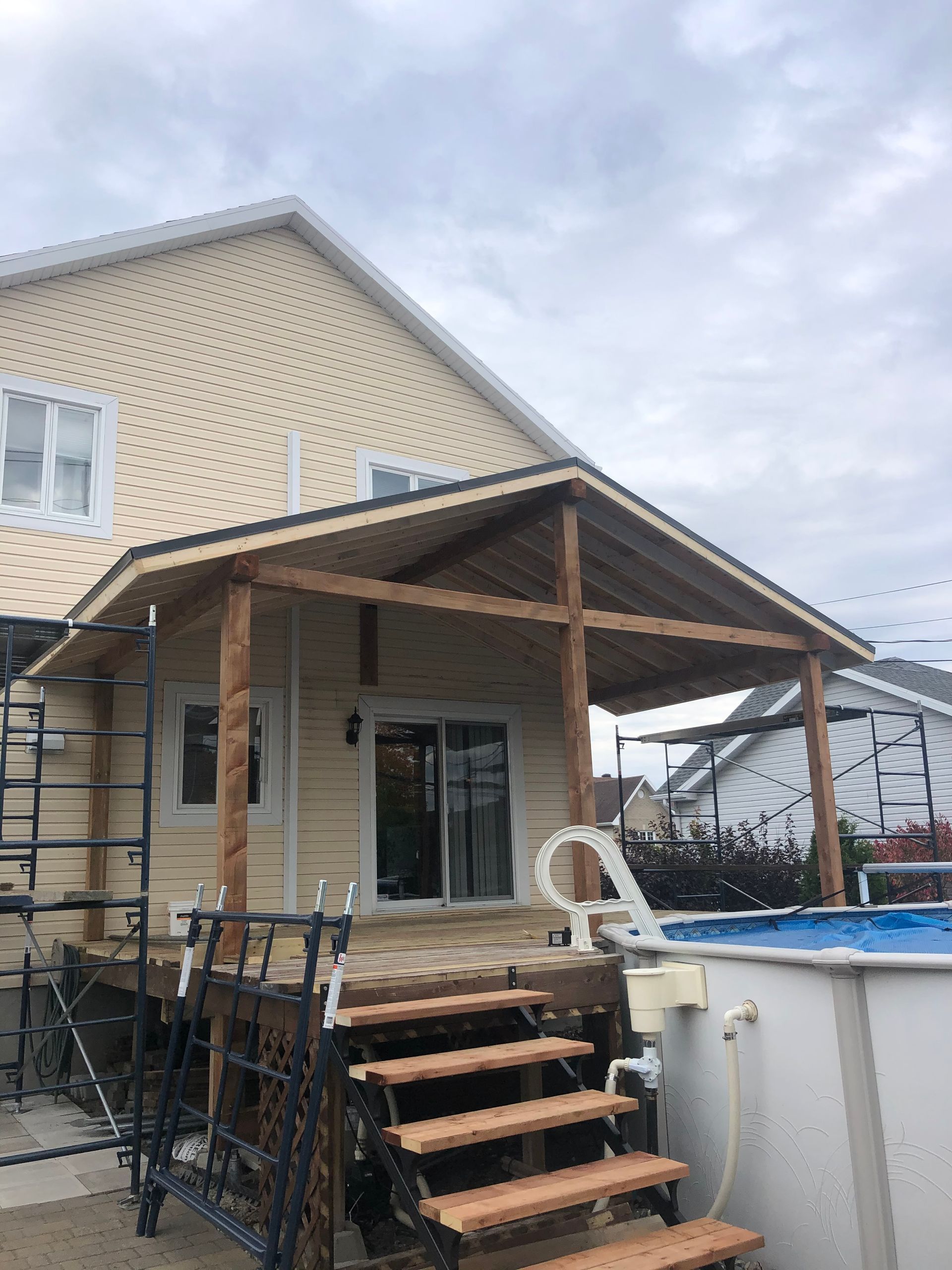 Maison beige avec terrasse en bois et patio couvert, piscine en arrière-plan.