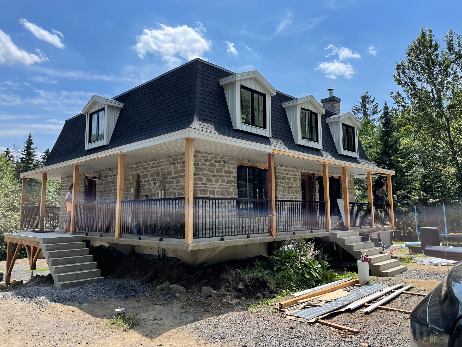 Maison revêtue de pierre, toit noir et véranda qui l'entoure. Des marches en pierre grise mènent à la véranda. Ciel bleu.