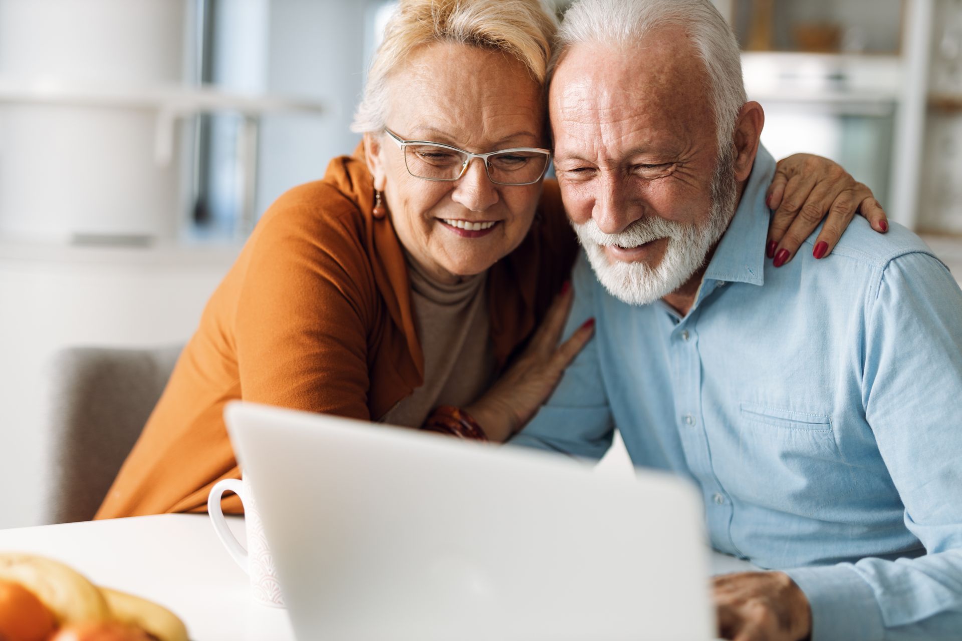 An elderly couple is sitting at a table looking at a laptop computer.
