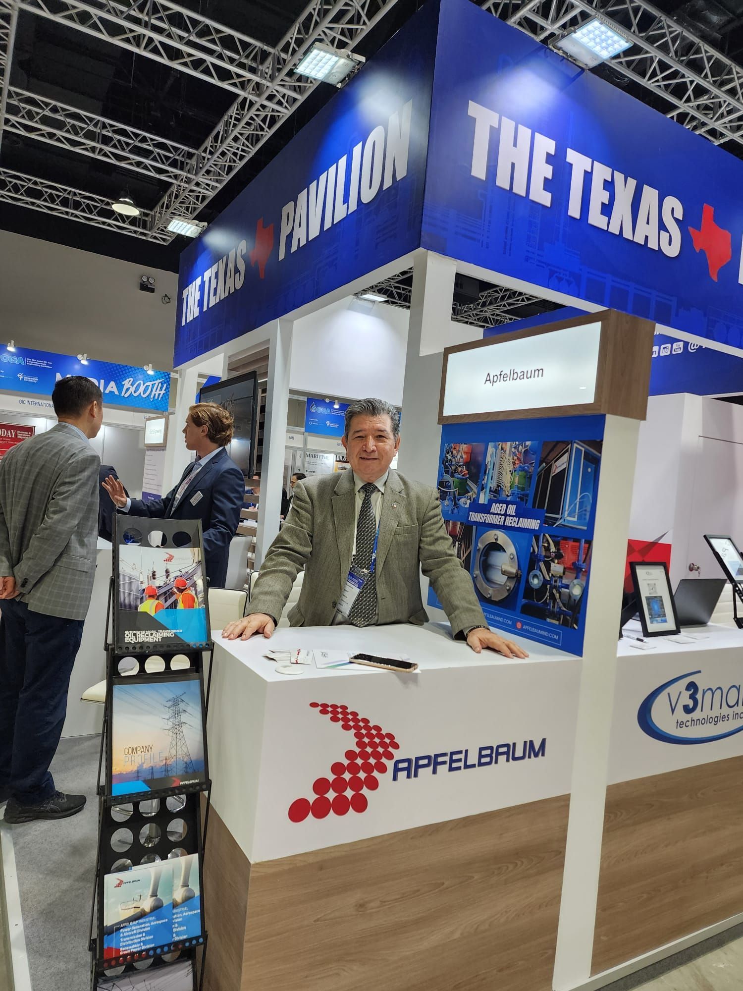 A person stands behind an Apfelbaum booth at a Texas Pavilion trade show, with another person visible in the background.