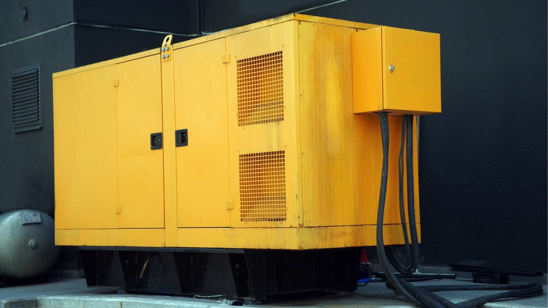 A yellow industrial diesel generator sits outside against a black wall, with electrical cables connected to its side.