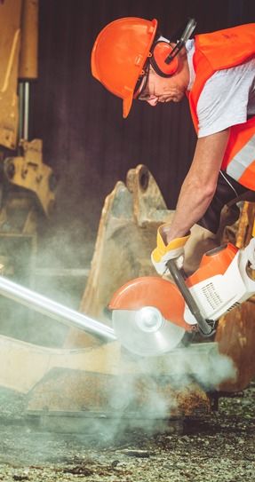 A construction worker in a high-visibility vest and hard hat uses a power saw to cut a metal pole, creating a cloud of dust.