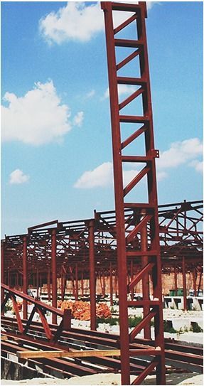 A tall, red steel lattice mast stands vertically on a construction site, with more metal framework visible in the distance.