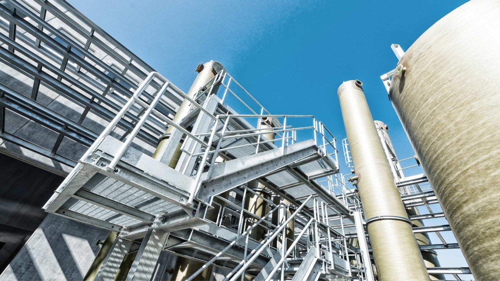 A low-angle view of industrial metal scaffolding, stairways, and cylindrical storage tanks against a clear blue sky.