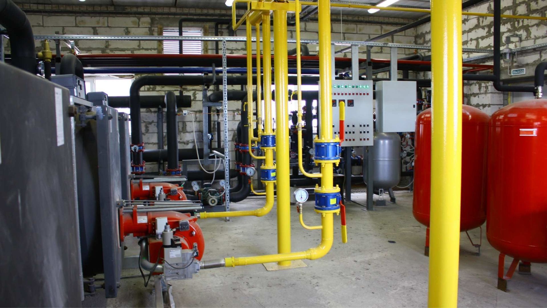 Industrial utility room featuring yellow gas pipes, control panels, and large red pressure tanks in a brick-walled space.