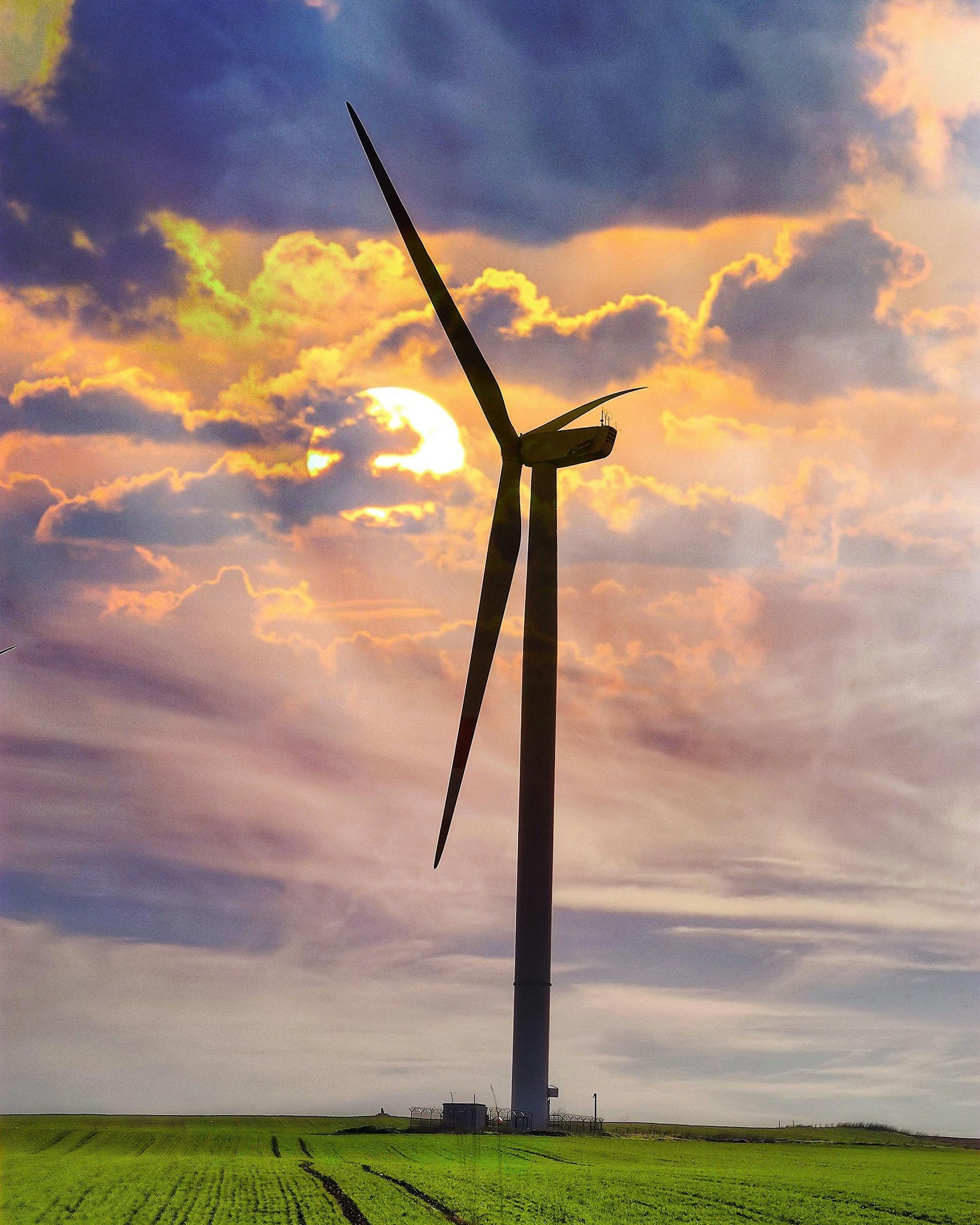 A wind turbine stands in a green field silhouetted against a cloudy sunset sky with golden light.