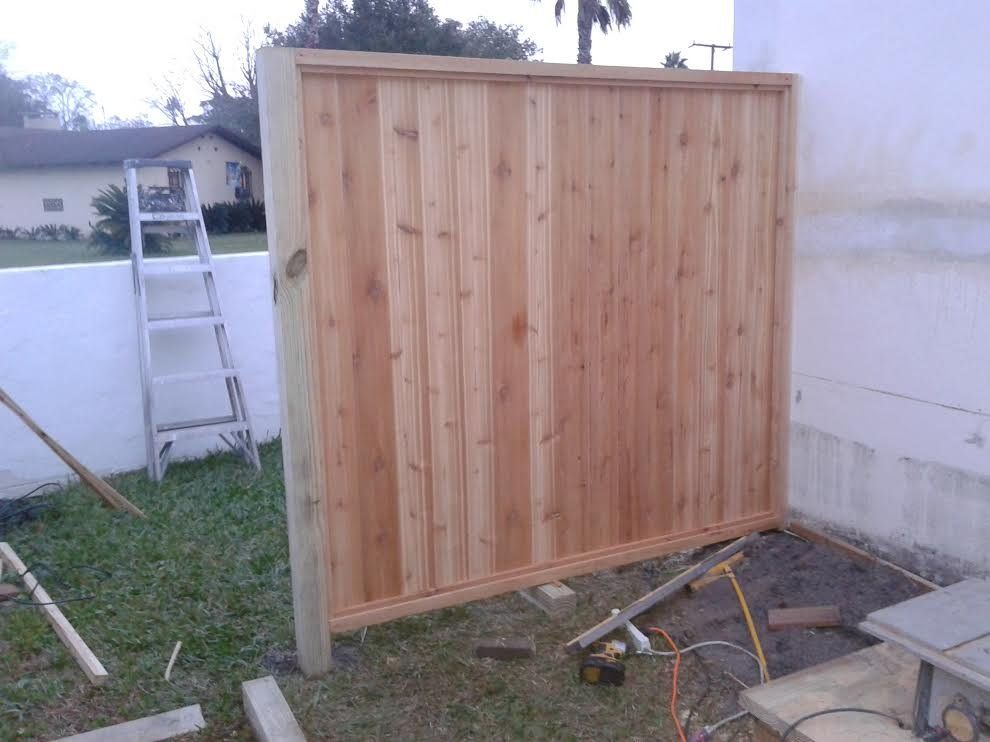 Wooden fence panel under construction beside a white wall, with a ladder and tools nearby.