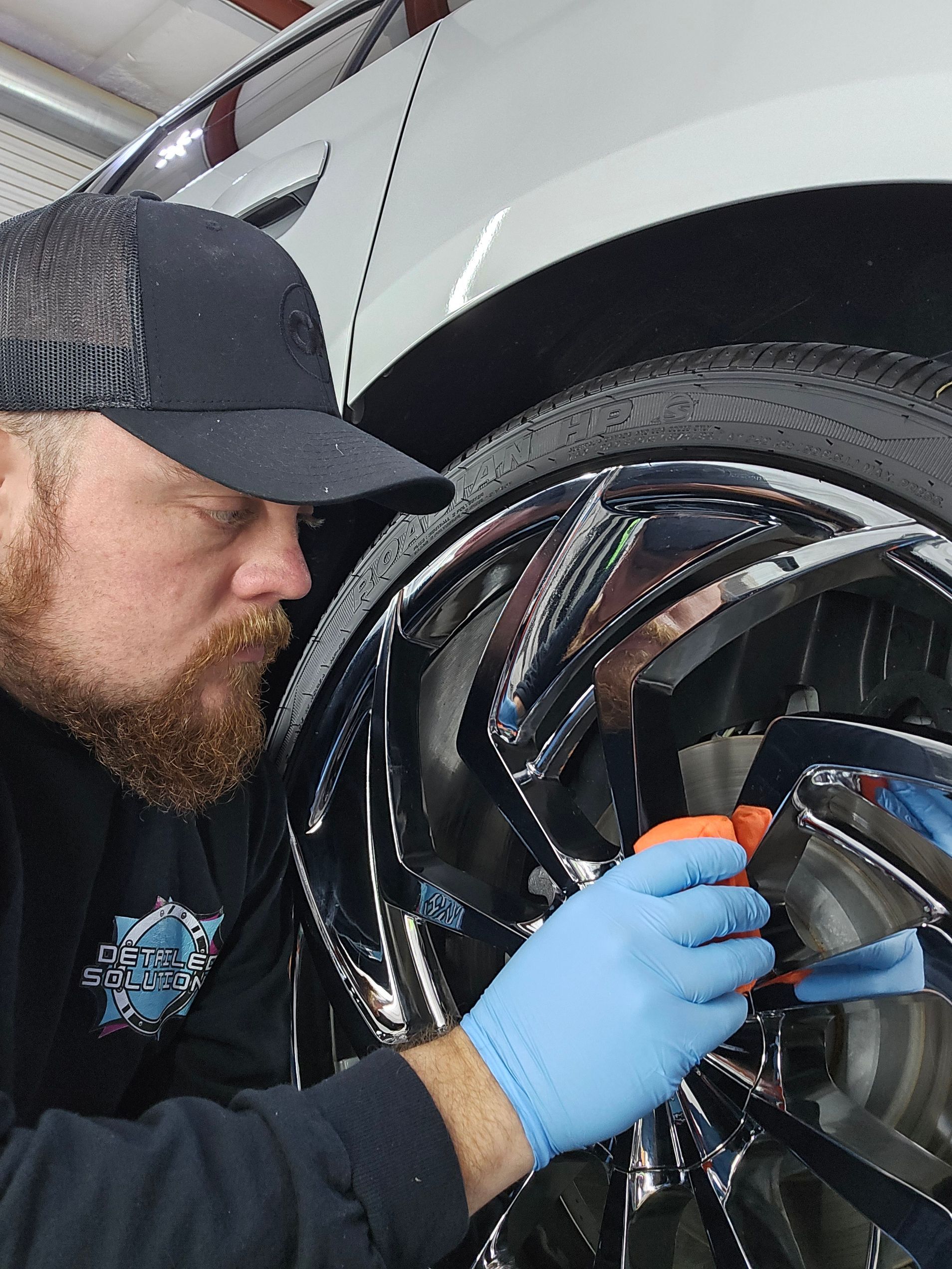 A person wearing a black hat and blue gloves cleans the chrome spokes of a car wheel.
