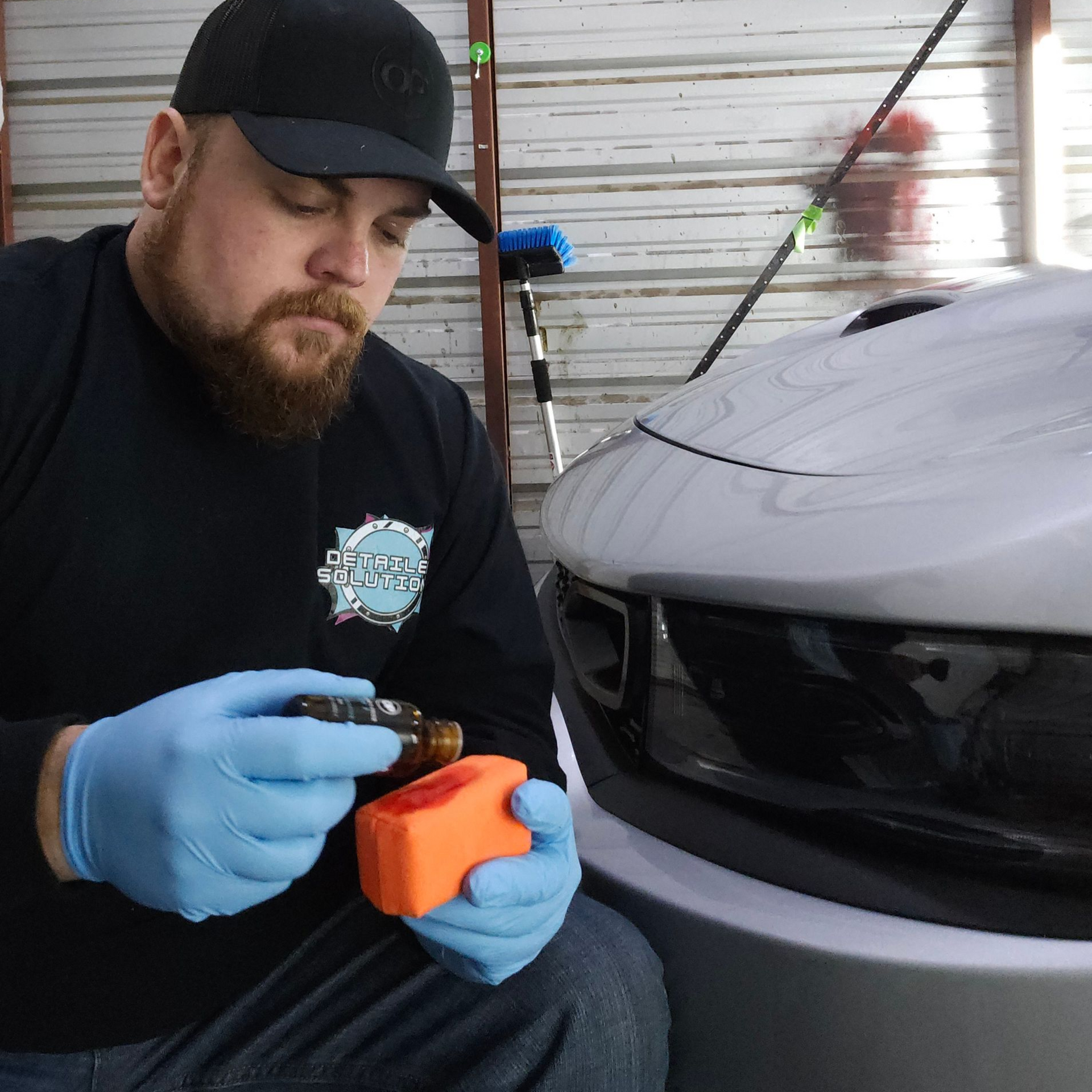 A person wearing blue gloves applies liquid from a small bottle onto an orange foam applicator near a car bumper.