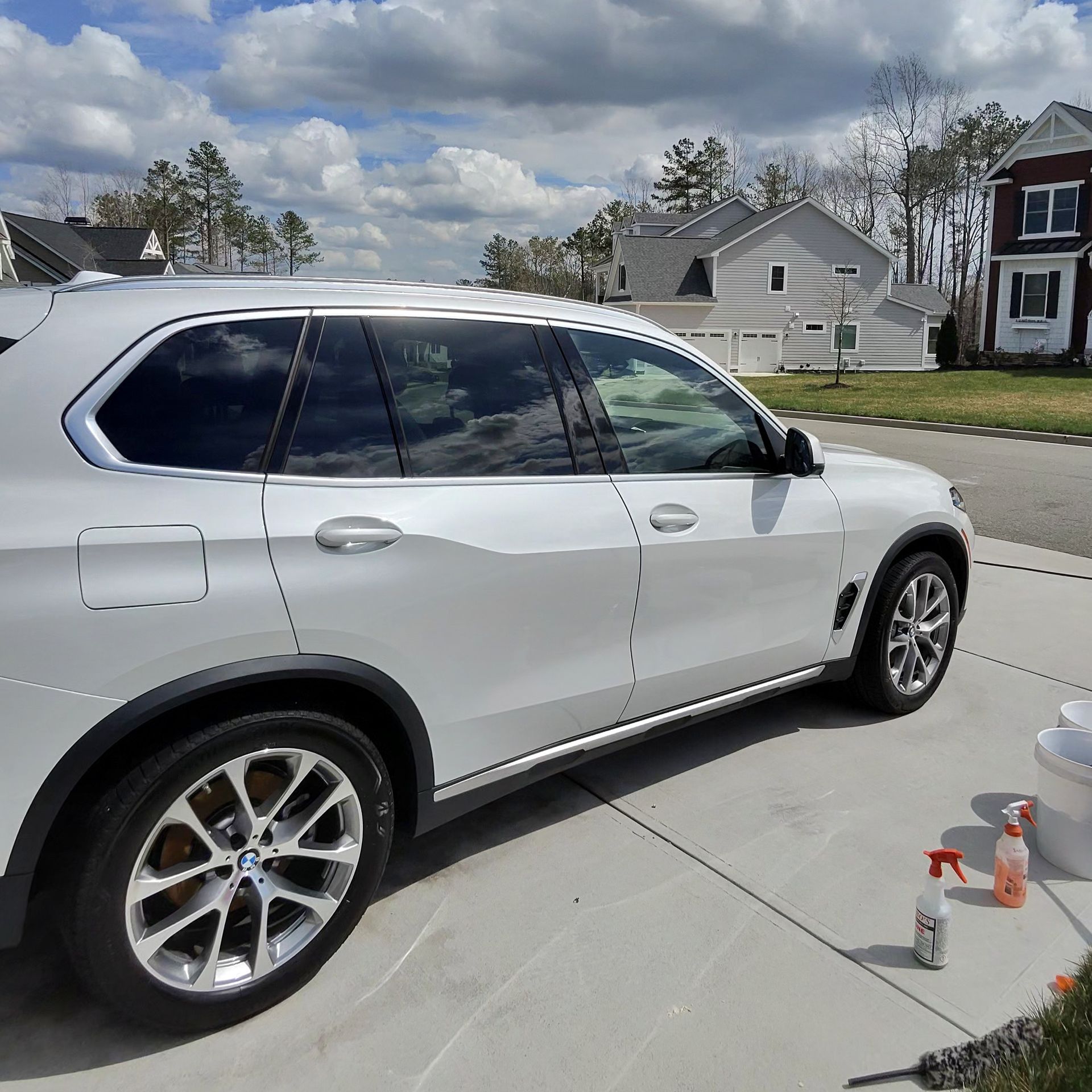 White SUV parked on a driveway with cleaning supplies; houses and sky in the background.