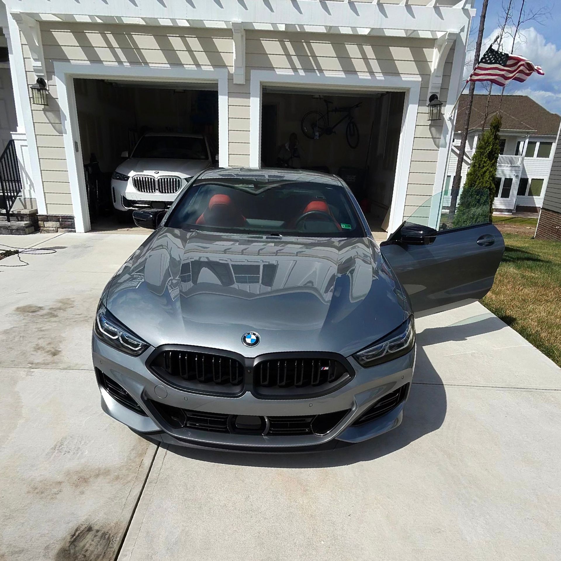 Gray BMW parked in front of a garage, driver's side door open. Another car visible in the garage.