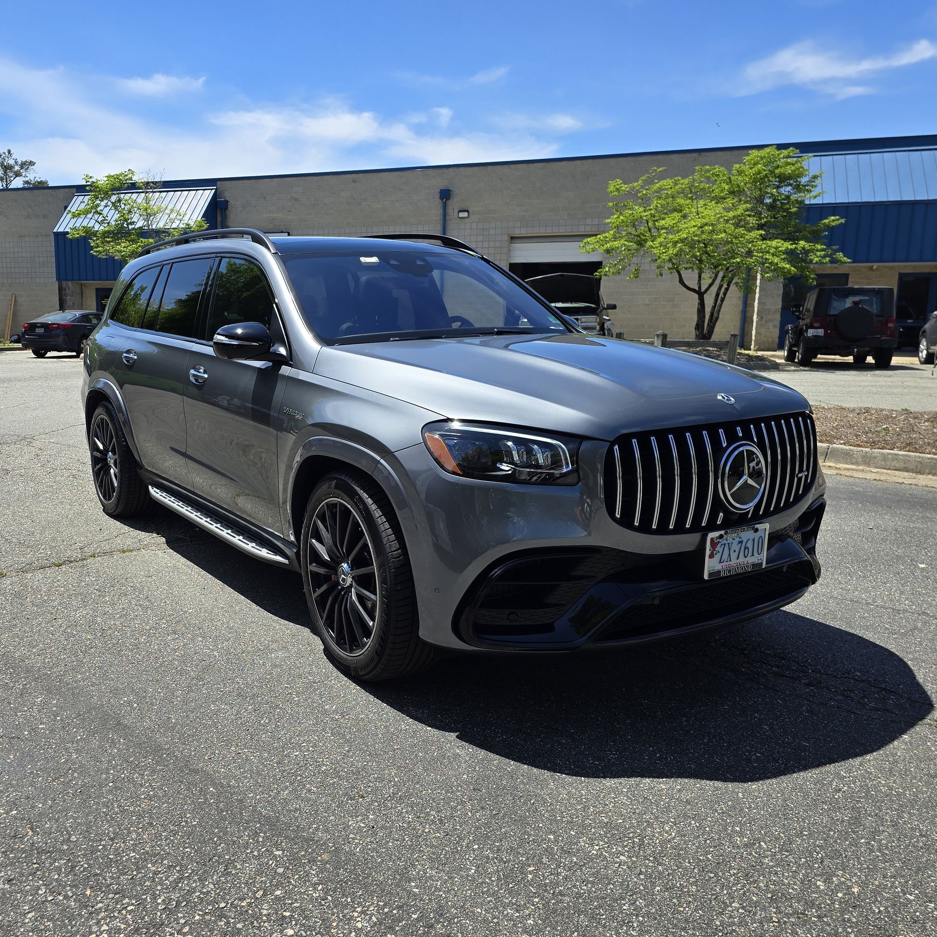 Gray Mercedes-Benz GLS SUV parked on pavement in front of a building with blue trim under a blue sky.