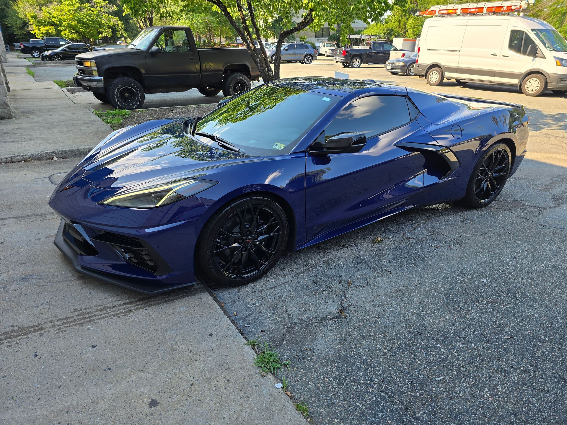 Blue Corvette sports car parked on a paved street, with a black pickup truck and other vehicles in the background.