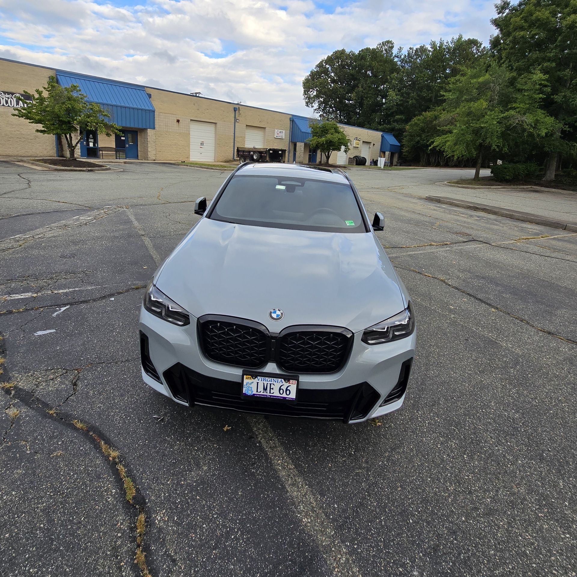 A gray BMW X3 M SUV parked in a paved lot, black grill, under a cloudy blue sky.