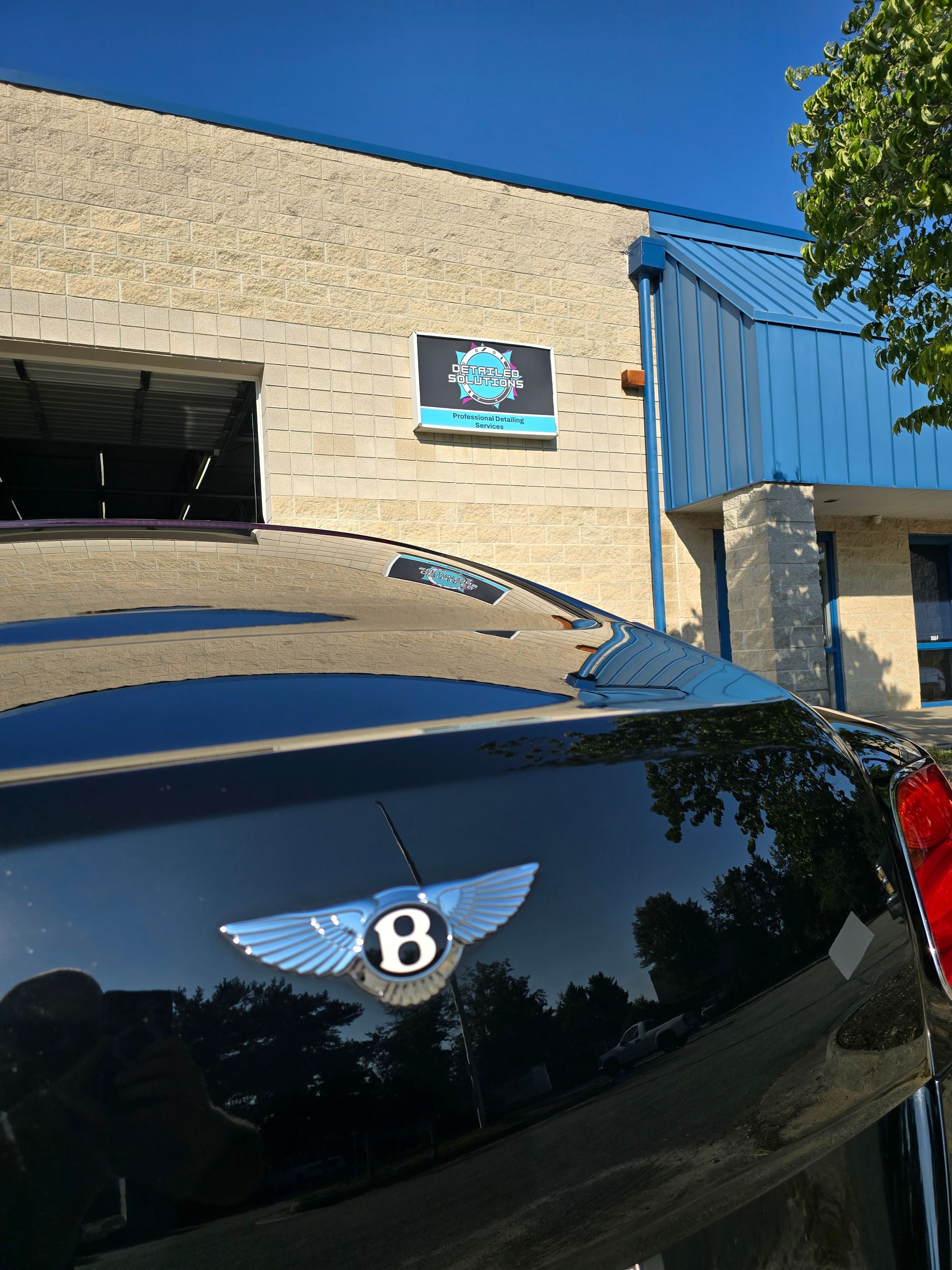 Black Bentley car with emblem, parked in front of a building with a blue sign.