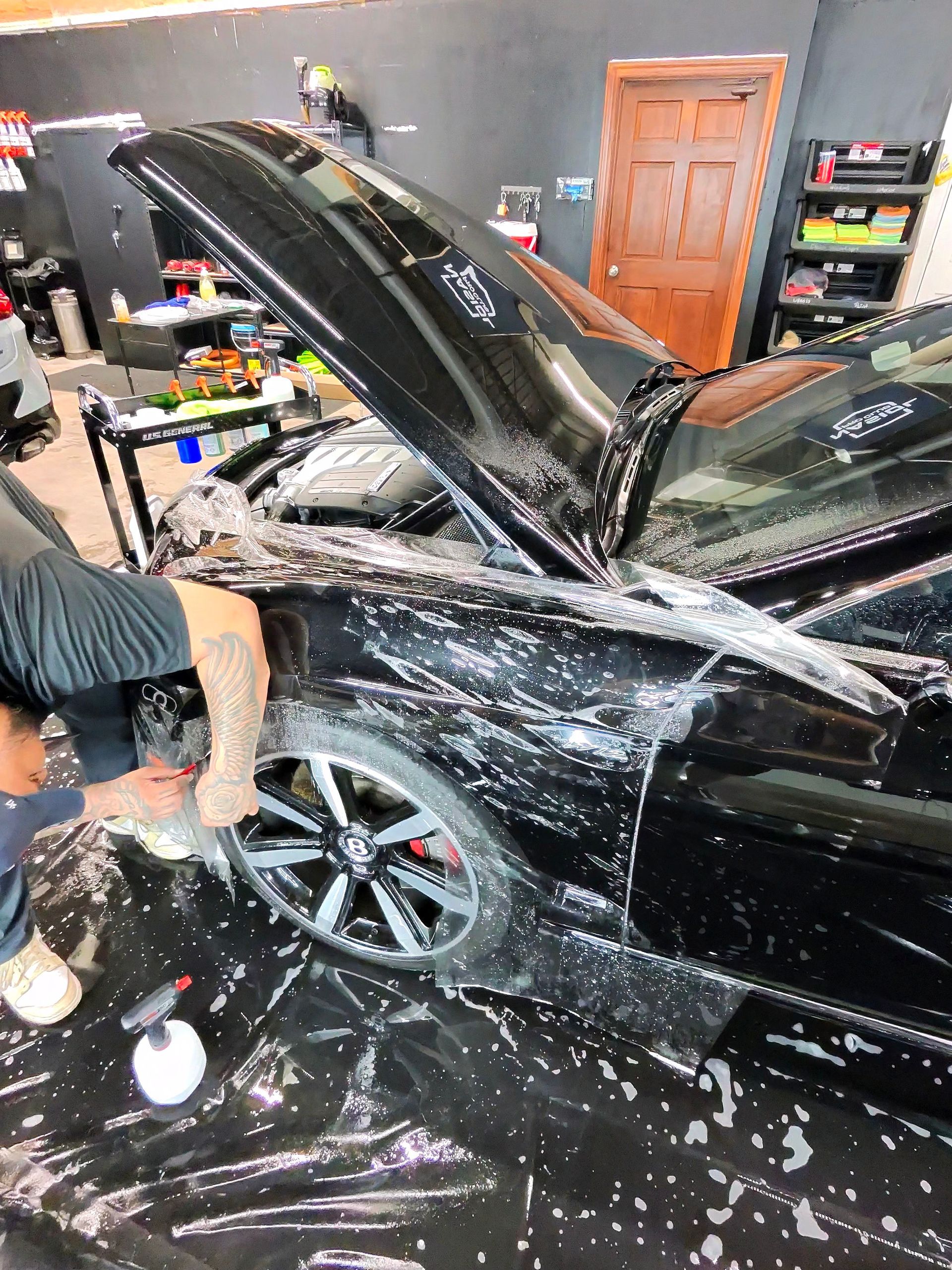 Man applying film to a black car in a garage; protective wrap covers the vehicle.