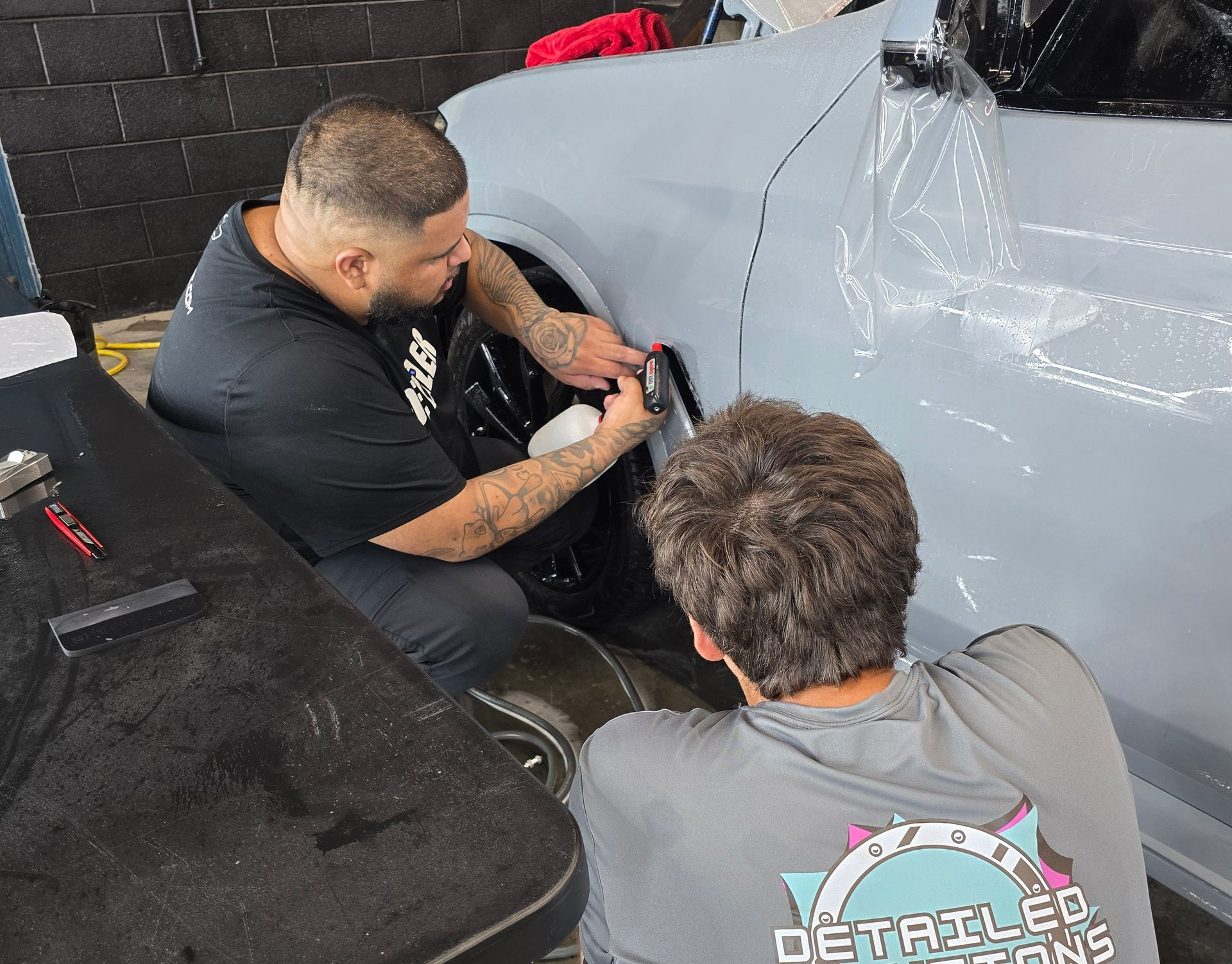Two people applying film to a light gray car. One sprays, the other smooths the film. Inside a shop.