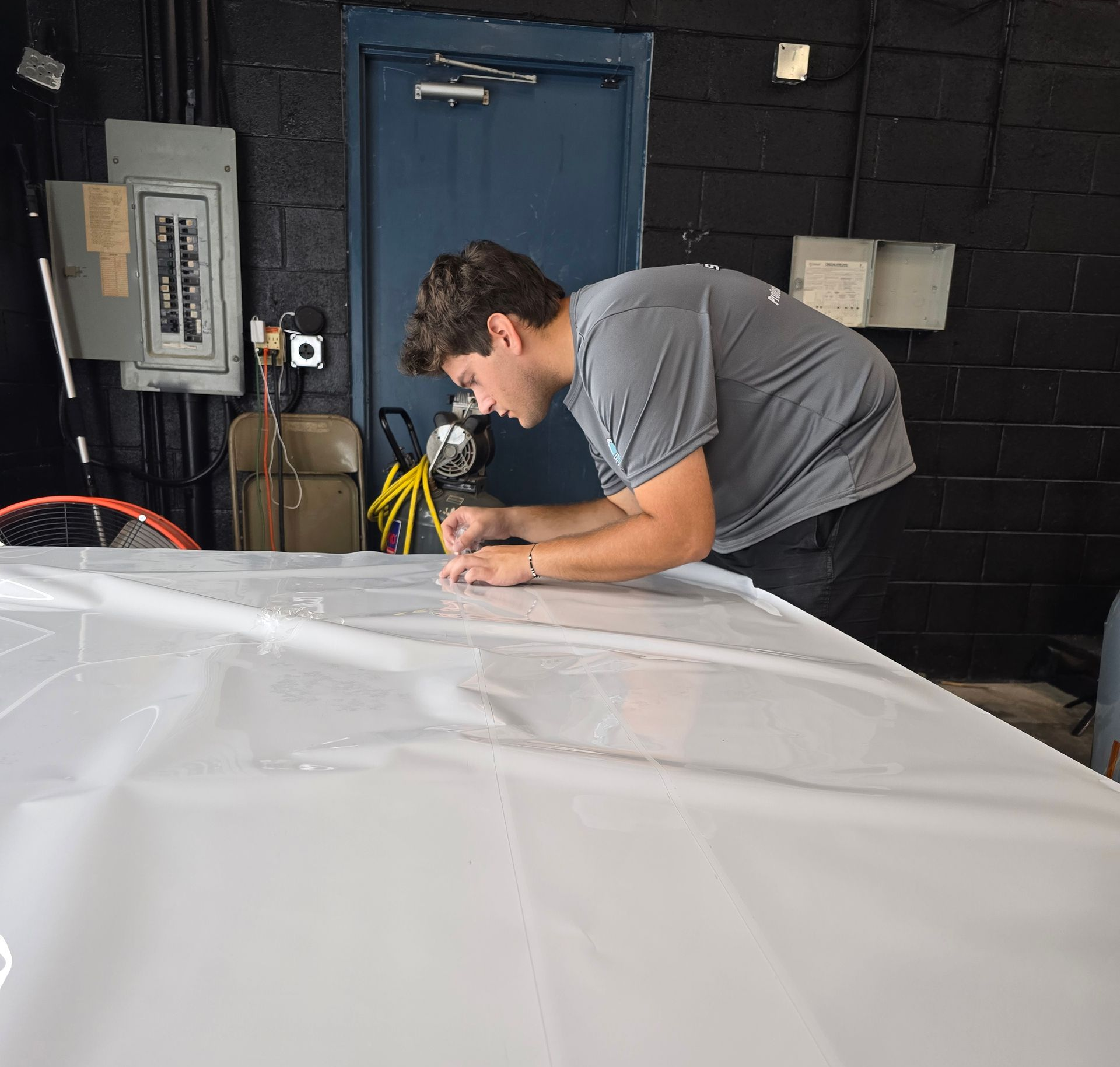 Man in gray shirt leans over a white surface, possibly drawing, in a workshop.