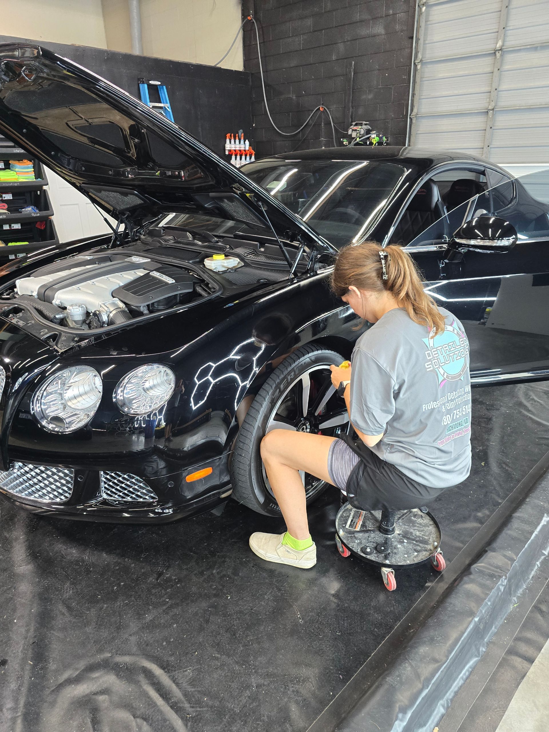 Person cleaning a black Bentley's wheel while seated on a rolling stool in a shop with the hood open.