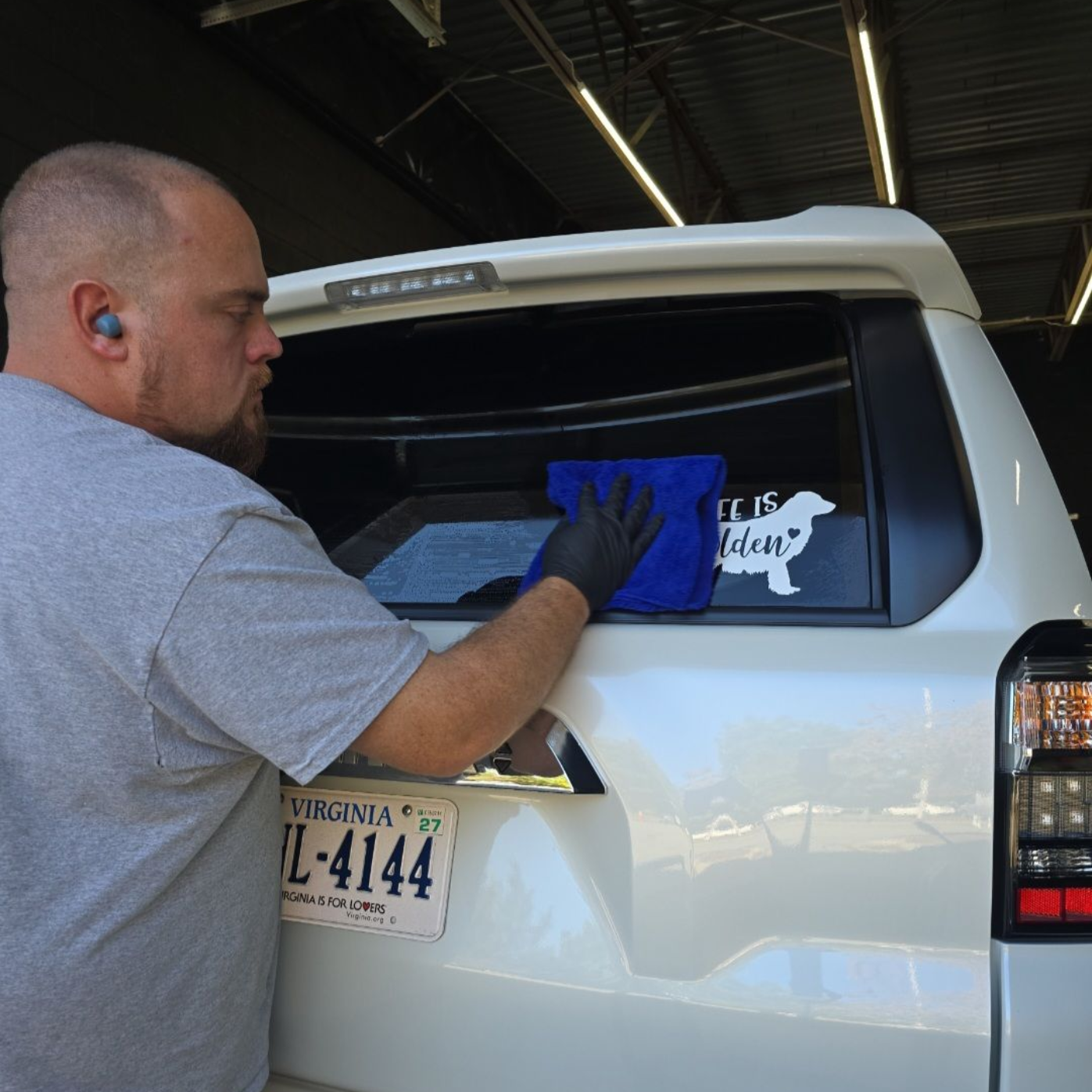 A person wearing a gray shirt and black glove cleans the rear window of a white SUV with a blue cloth in a garage.