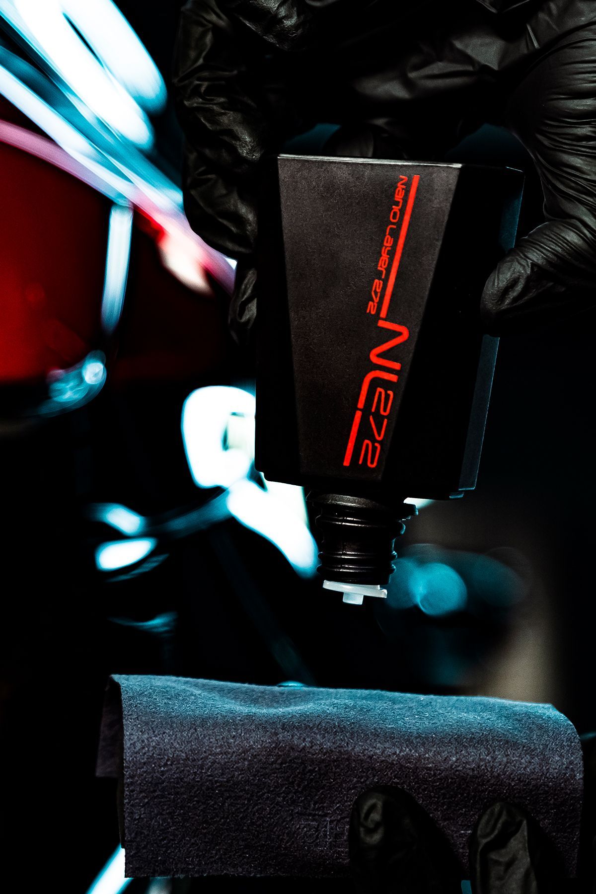 Gloved hands dispensing detailing product onto a microfiber cloth in front of a car. Black and red product packaging.