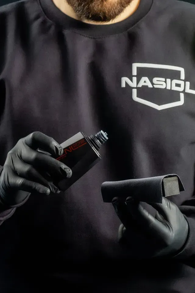 A person in a Nasiol branded black shirt and gloves applying a protective liquid from a bottle onto a foam applicator block.