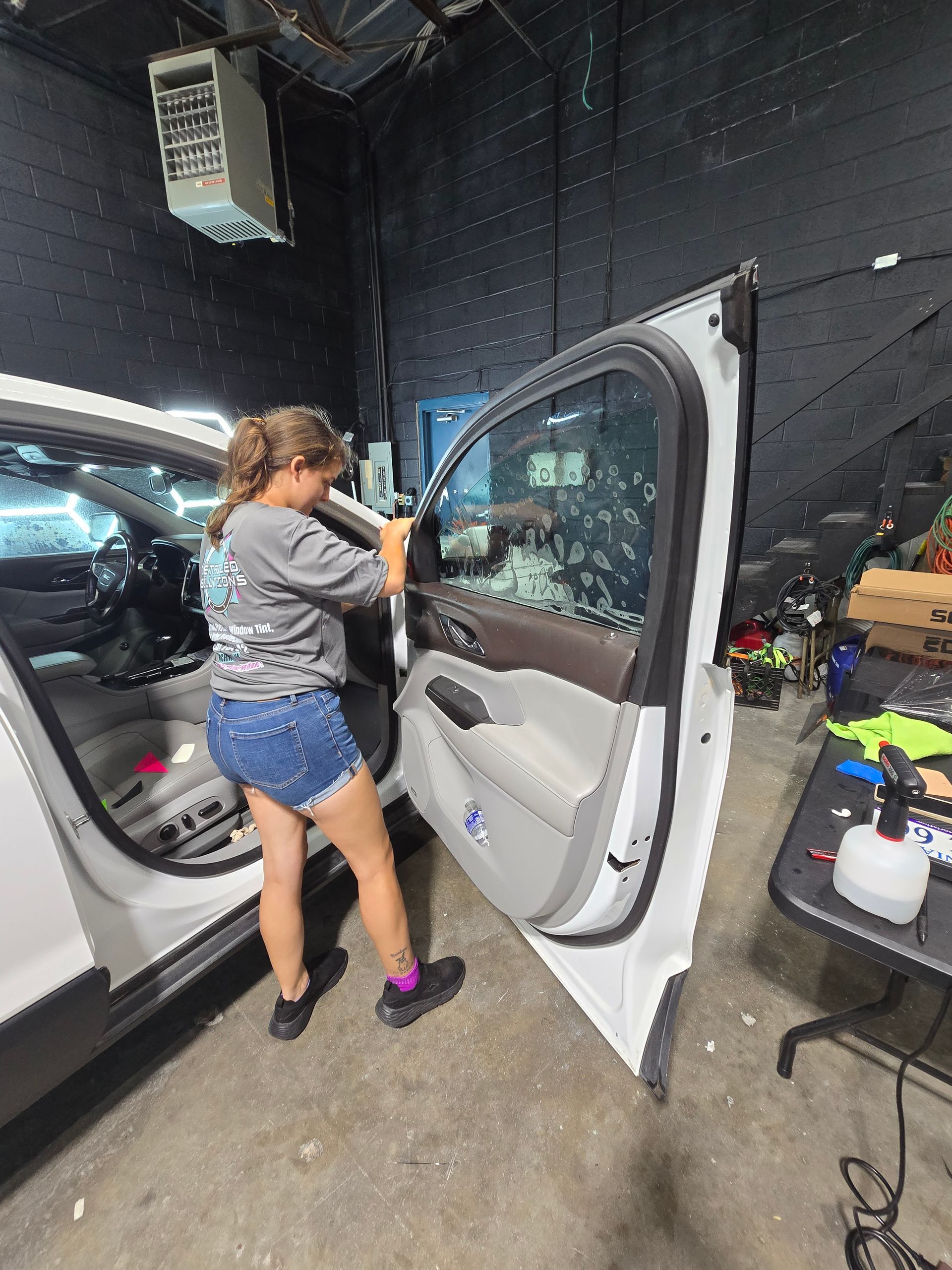 Woman applying tint to a car window inside a garage. She is wearing shorts and a t-shirt.