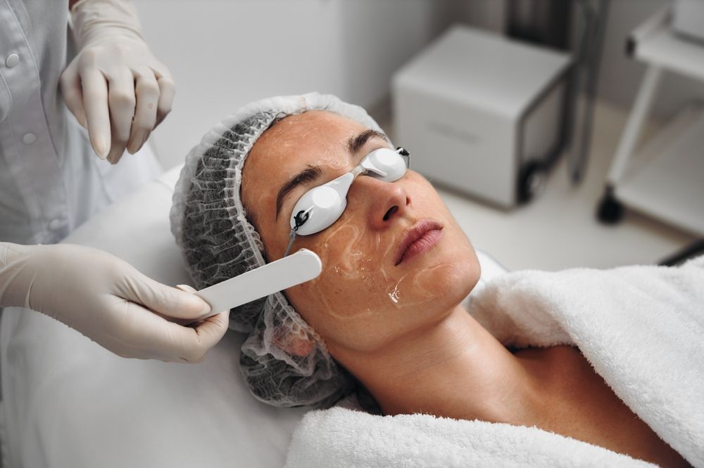 Woman undergoing facial laser treatment at a spa, wearing protective eyewear and cap. A gloved hand holds a spatula applying gel to her face.