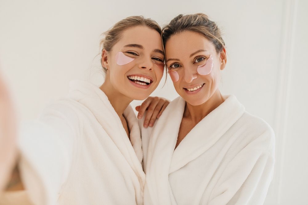 Two people in white bathrobes wearing pink under-eye patches, smiling at the camera during a skincare routine.