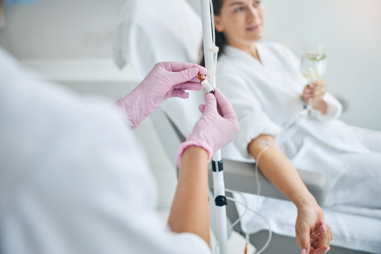 A healthcare worker in pink gloves adjusting an IV drip for a woman in a white robe, indoors.