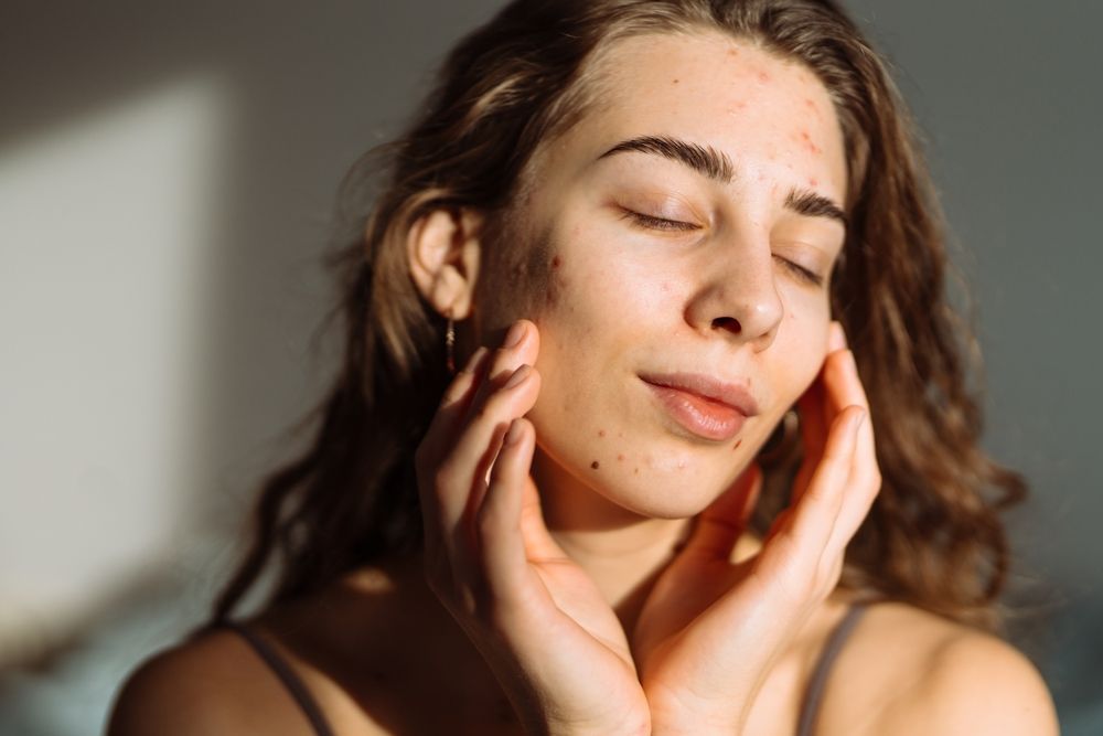 A person with natural skin texture gently touches their face with both hands, eyes closed, in soft, warm side-lighting.