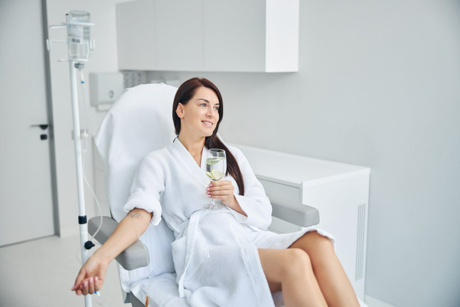 Woman in robe receiving IV drip, holding drink, relaxing in spa-like setting.
