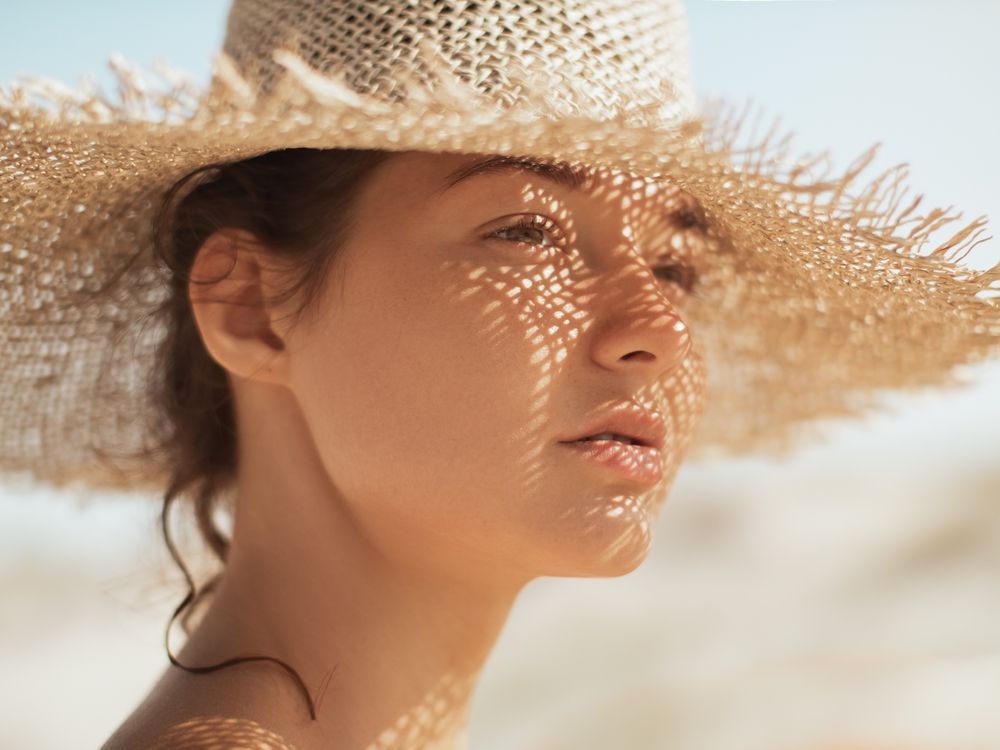 A person wearing a wide-brimmed straw hat with sunlight casting a textured, cross-hatched shadow across their face.