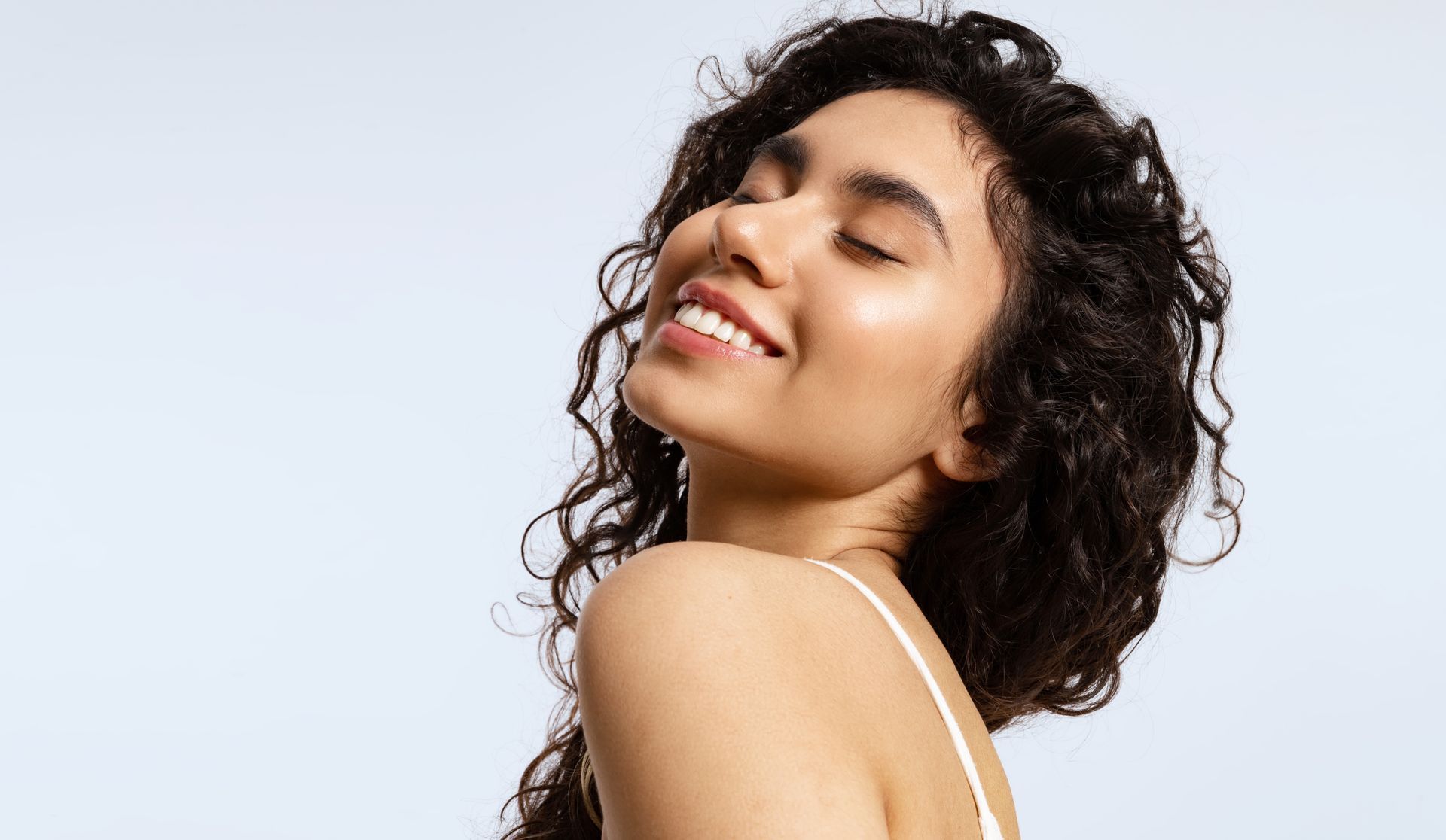 Woman with curly dark hair smiles with eyes closed, facing upward, against a light blue backdrop.