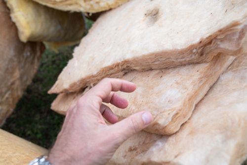 Hand touching a stack of light brown insulation batts outdoors.