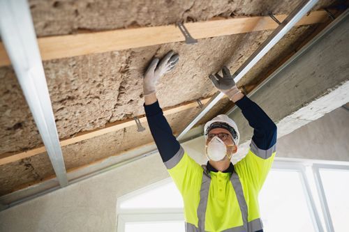 Construction worker installing insulation in ceiling, wearing safety gear: mask, gloves, high-vis vest.