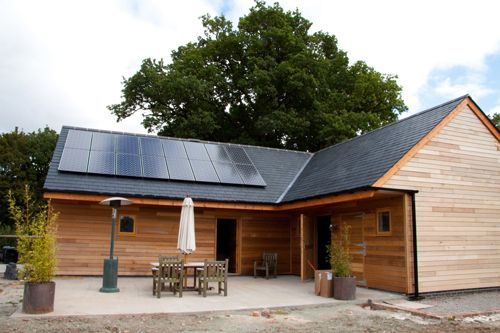 Wooden building with solar panels, outdoor seating, and a large tree in the background.