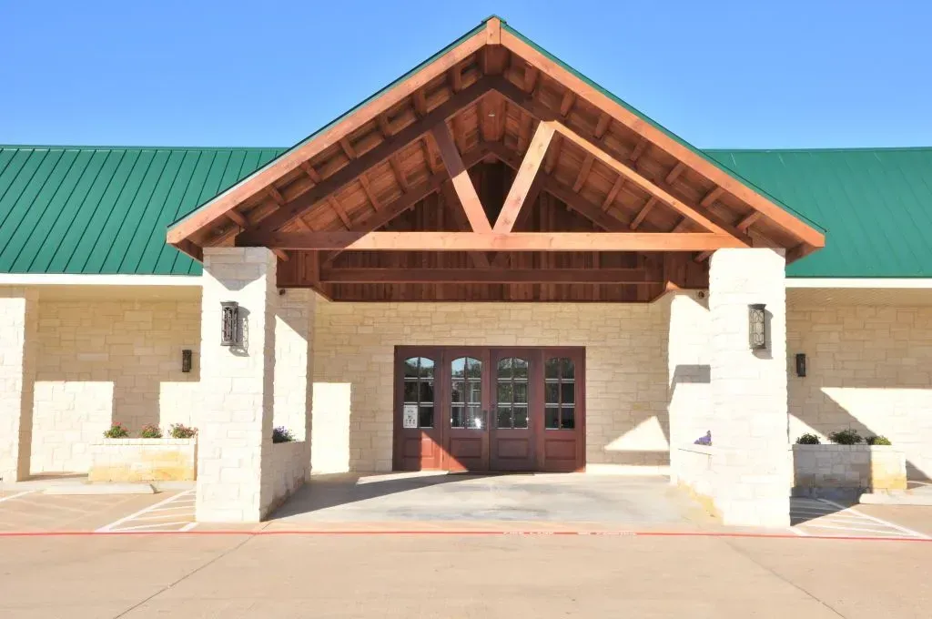 A single-story building with a tan stone facade, green roof, and a prominent timber-framed covered entrance.