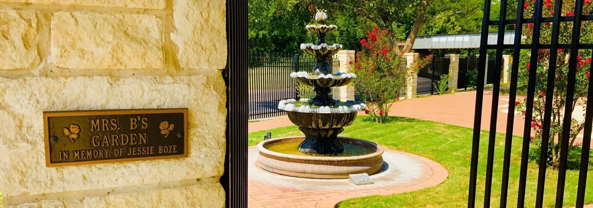 A three-tiered fountain stands in a garden viewed through an open gate, with a plaque on the stone wall nearby.