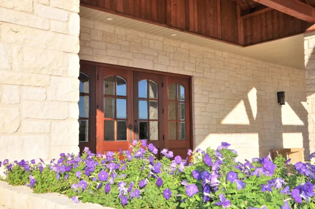 A patch of vibrant purple petunias in front of a stone building entrance with dark wooden double doors.
