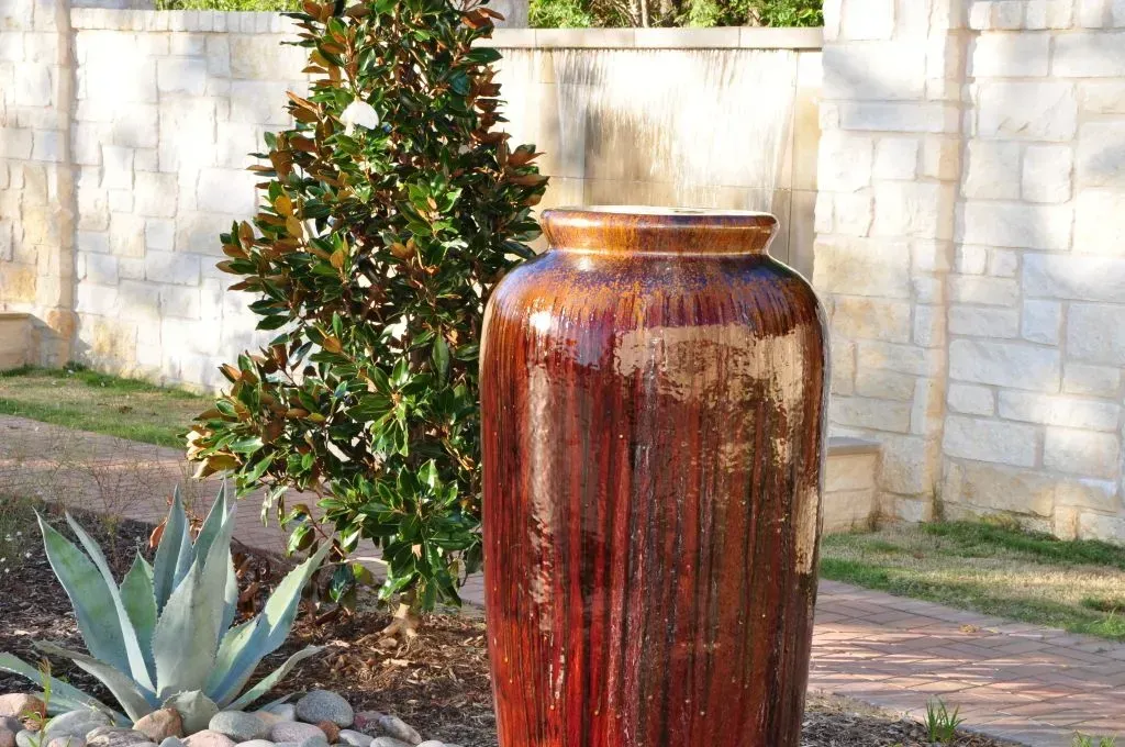A large, glazed, brown ceramic urn standing in a garden next to a small tree and an agave plant against a stone wall.