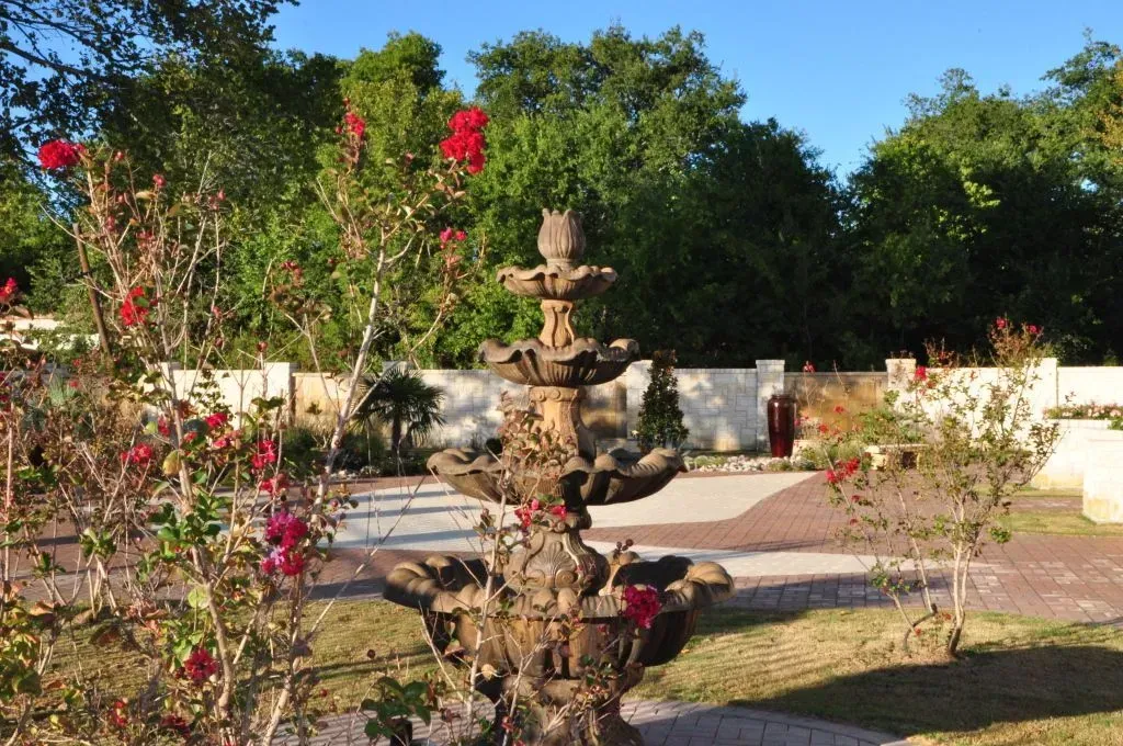 A tiered stone fountain surrounded by rose bushes and a paved patio under a clear blue sky.