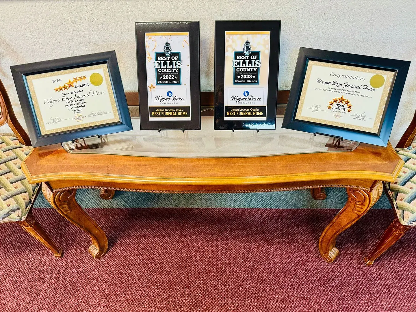 Four framed awards displayed on a wooden table between two chairs on a patterned carpet.