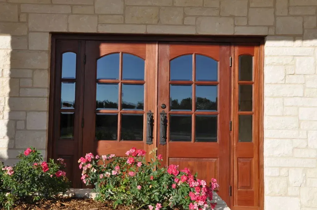 A wooden double door with glass panels and sidelights set in a stone wall, with pink flowers in the foreground.