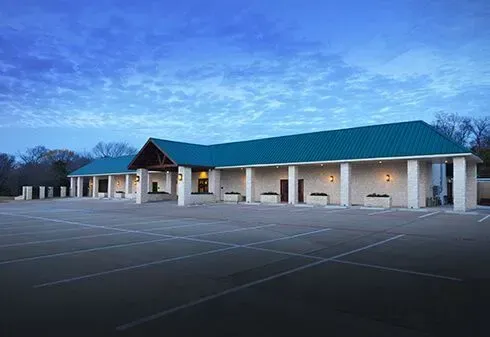 A single-story commercial building with a teal metal roof and a stone facade, fronted by an empty asphalt parking lot.