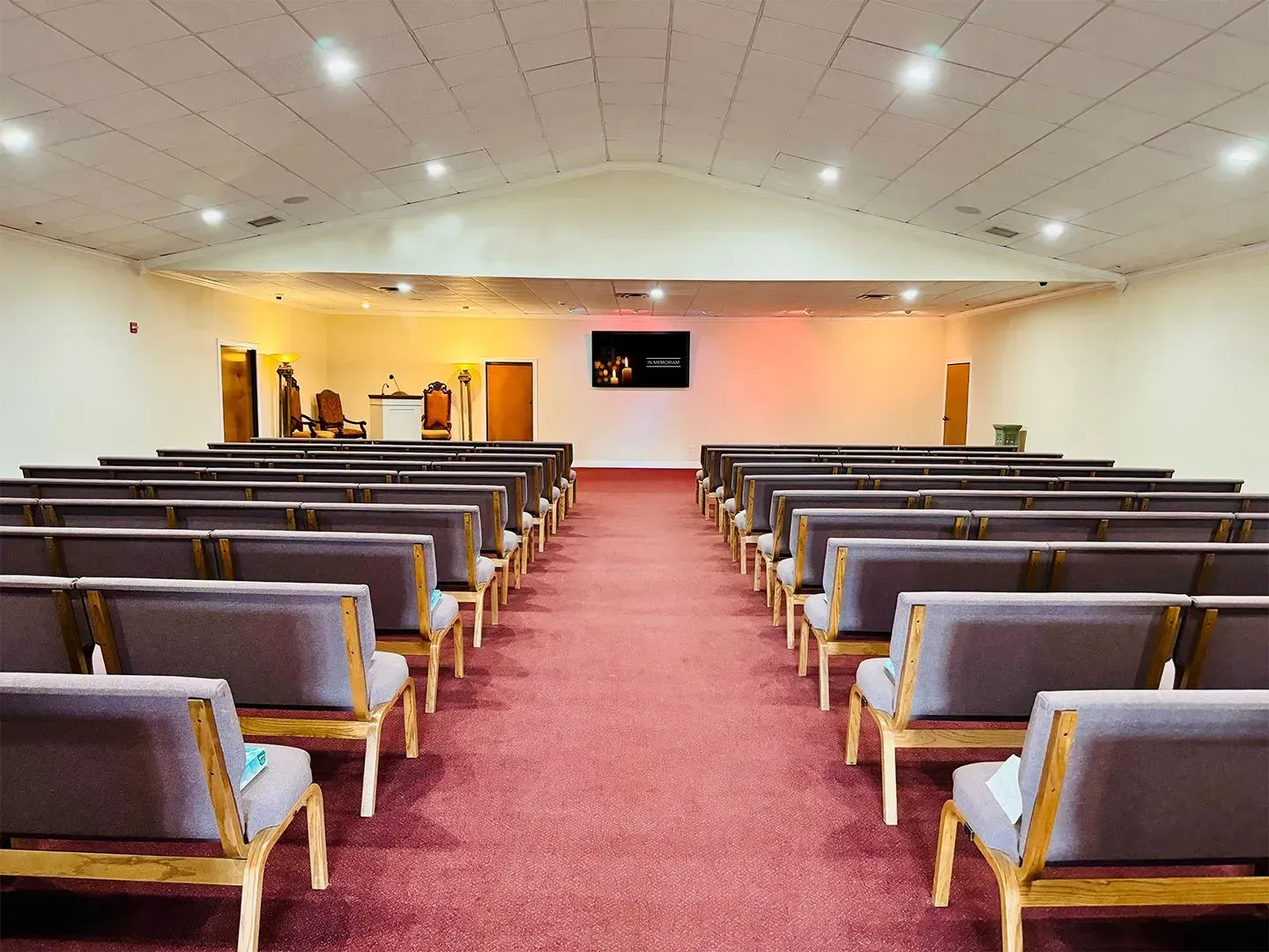 Empty church sanctuary with rows of grey padded pews facing a stage with a screen and warm lighting on a red carpet.